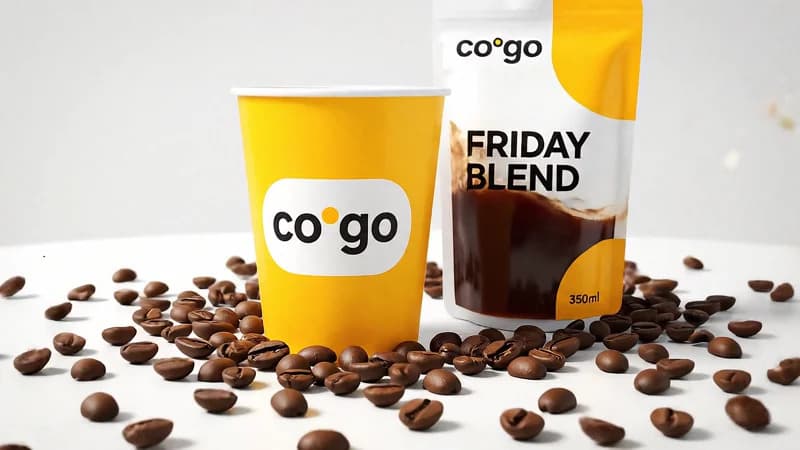 A yellow CoGo takeaway cup next to a Friday Blend pouch on a bed of coffee beans